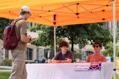 Two students sit at a folding table beneath an orange tent. A third student approaches the tent.