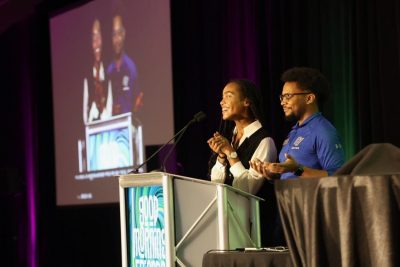 A young woman stands at a lectern, smiling and speaking in a dark conference room with a projection screen in the background. A young man stands with her, smiling and gesturing.