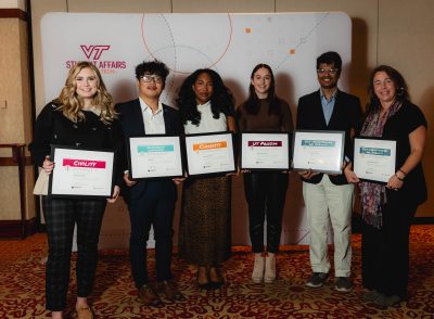 Six smiling people stand shoulder-to-shoulder and hold their variously colored award certificates.